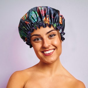 May include: A woman with a bright smile is wearing a black satin shower cap with a colorful floral print. The cap has a ruffled edge.