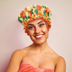 May include: A yellow shower cap with an orange and green citrus fruit pattern. The cap has an orange ruffled trim. The person is wrapped in a coral towel.