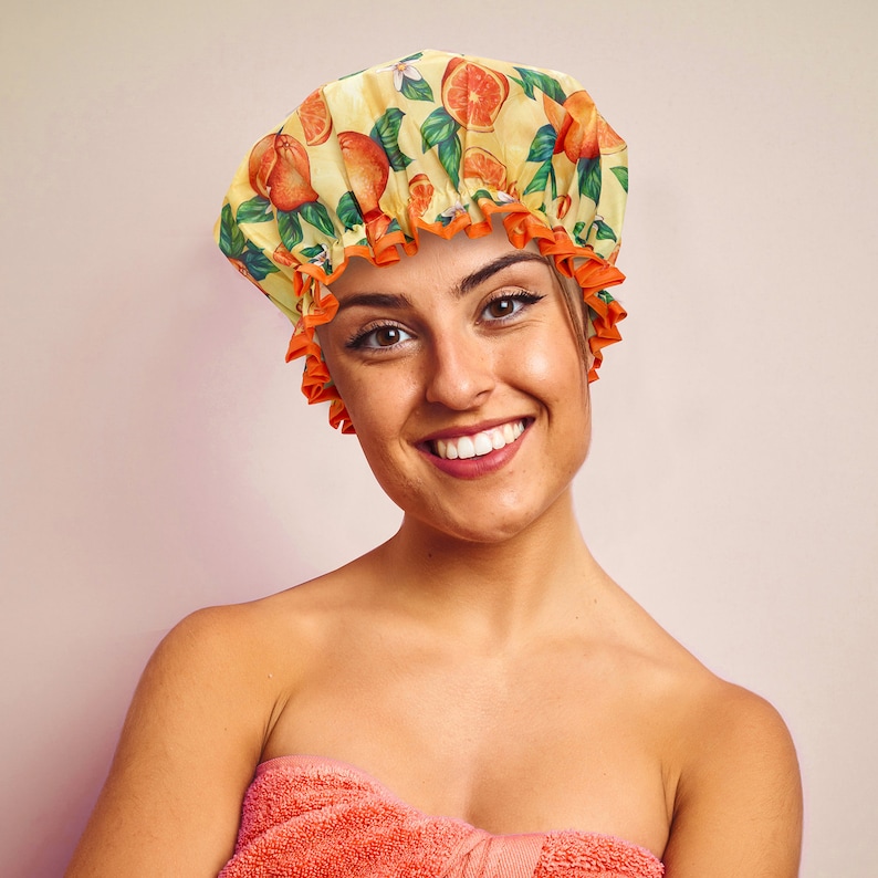 May include: A yellow shower cap with an orange and green citrus fruit pattern. The cap has an orange ruffled trim. The person is wrapped in a coral towel.