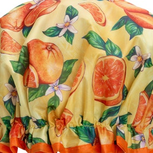May include: A yellow shower cap with a vibrant orange and white floral pattern. The design features oranges, orange slices, white flowers, and green leaves. The cap has an orange trim.
