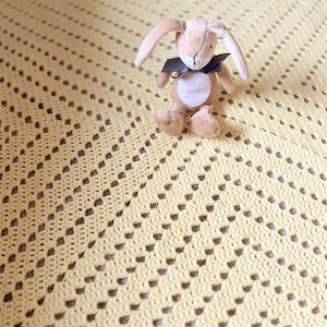 May include: A light yellow crocheted baby blanket with a repeating openwork pattern. A small stuffed bunny with a black bow sits on the blanket. The blanket is made of soft yarn and is suitable for a baby.