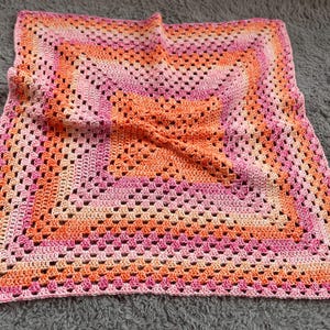 Pode incluir: Um cobertor quadrado de croch&ecirc; feito &agrave; m&atilde;o em tons de rosa e laranja. O cobertor apresenta um design de granny square com um padr&atilde;o repetitivo de quadrados vazados. As cores transitam do rosa claro para o laranja em quadrados conc&ecirc;ntricos.