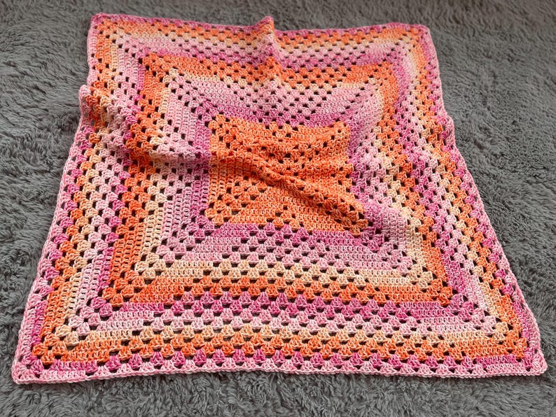 Pode incluir: Um cobertor quadrado de croch&ecirc; feito &agrave; m&atilde;o em tons de rosa e laranja. O cobertor apresenta um design de granny square com um padr&atilde;o repetitivo de quadrados vazados. As cores transitam do rosa claro para o laranja em quadrados conc&ecirc;ntricos.