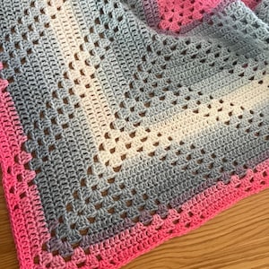 Könnte beinhalten: Eine handgefertigte Häkeldecke mit Farbverlauf. Die Decke geht von Rosa über Grau und Weiß zu einem hellrosa Rand über. Das offene Webmuster verleiht dieser kuscheligen, weichen Decke Textur und visuelles Interesse.