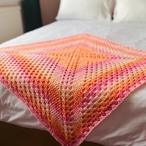 May include: A square crochet blanket featuring a vibrant gradient pattern of pink, orange, and yellow yarn. The design is constructed with a classic granny square stitch pattern, radiating outward from the center in concentric diamond-shaped layers.