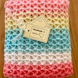 Op de afbeelding: Een handgemaakte, veelkleurige gehaakte deken in roze, gele, blauwe en witte tinten. De deken is vastgebonden met een transparant lint en bevat een label met de tekst "Handmade with love."