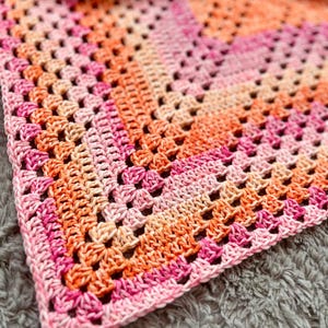 Op de afbeelding: Een handgemaakte gehaakte deken in tinten roze, oranje en perzik. De deken heeft een granny square design met open vierkanten en een effen rand. De garenkleuren vloeien in elkaar over, wat een warme en uitnodigende uitstraling creëert.