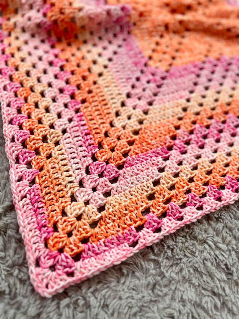 Pode incluir: Um cobertor de croch&ecirc; feito &agrave; m&atilde;o com um padr&atilde;o de quadrados da vov&oacute;. O cobertor apresenta um gradiente de cores, que vai do rosa claro ao laranja. O design de tecido aberto e a textura macia sugerem um item aconchegante.