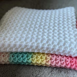 May include: A stack of two crocheted blankets. The top blanket is white, with a textured, honeycomb-like pattern. The bottom blanket has a rainbow gradient border of pink, yellow, green, and white.