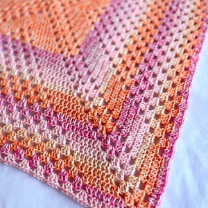 May include: A square crochet blanket featuring a gradient color pattern that transitions through shades of orange, coral, and pink. The textile is constructed with a classic granny square stitch pattern, creating a textured, open-weave design.