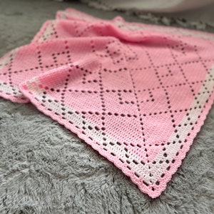 Könnte beinhalten: Eine rosa gehäkelte Babydecke mit einem weißen gehäkelten Rand. Die Decke hat ein Rautenmuster.