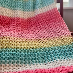 Op de afbeelding: Een handgemaakte gehaakte deken met een gestructureerd steekpatroon. De deken heeft horizontale strepen in pastelkleuren: roze, geel, turquoise en wit. De deken is over een houten stoel gedrapeerd en toont de zachte textuur en kleurovergang.