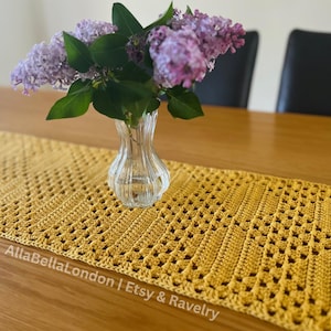 May include: A handmade, golden yellow crochet table runner with a geometric openwork design. A clear glass vase holds a bouquet of purple lilacs. The table runner is on a wooden table.