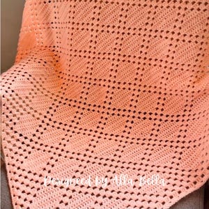 May include: A peach-colored crochet blanket featuring a repeating diamond pattern created with openwork stitches. The fabric has a textured, grid-like appearance throughout. The text "Designed by Alla Bella" is printed in white across the lower section.