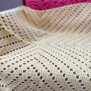 Könnte beinhalten: Eine cremefarbene, handgefertigte Häkeldecke mit einem sich wiederholenden Lochmuster. Die Decke ist gefaltet und zeigt die Textur und das Design. Die Häkelarbeit wirkt weich und zart, geeignet für eine Babydecke oder einen dekorativen Überwurf.