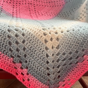 May include: A handmade crochet blanket in shades of pink, gray, and white. The blanket features a granny square design with a gradient effect, transitioning from pink to gray and white. The blanket is folded over a chair.