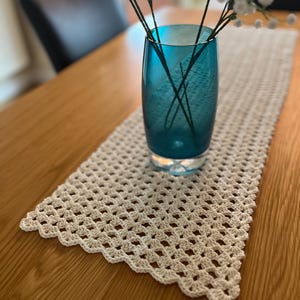 May include: A teal glass vase holding white flowers sits on a cream-colored, crocheted table runner. The runner has a scalloped edge and is placed on a wooden table. The vase is cylindrical and the flowers have delicate white blossoms.