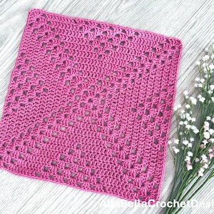 May include: A magenta crochet square with an openwork pattern. The handmade item is displayed on a light grey wooden surface, with a small bunch of white flowers in the background. The crochet piece is likely a washcloth or decorative square.