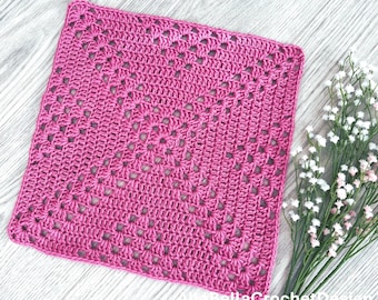 May include: A square, magenta-colored crochet piece with an openwork design. The handmade item is placed on a light gray wooden surface, with a small bouquet of white flowers in the background. The crochet piece is likely a washcloth or a decorative square.