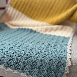 Puede incluir: Una manta de ganchillo hecha a mano en tonos de verde azulado, crema y amarillo mostaza. La manta presenta un patrón de puntadas texturizadas y un borde festoneado. La manta está sobre una silla.