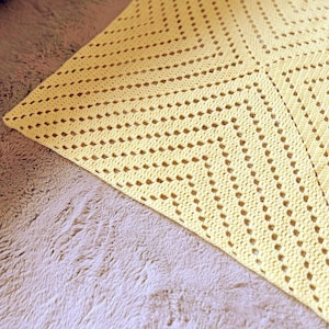 May include: A handmade, yellow crochet blanket with a geometric pattern. The blanket is made of soft yarn and features a repeating design of openwork stitches, creating a textured and airy feel. The blanket is folded, showing its corner.