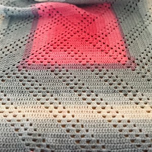 May include: A handmade crochet blanket with a gradient design. The blanket transitions from white to gray, with a central pink square. The crochet pattern features openwork details, creating a textured appearance. The blanket is likely made of soft yarn.