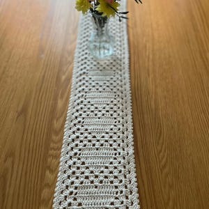 May include: A cream-colored, rectangular crochet table runner with an openwork design. A small glass vase holds pink and yellow flowers, placed in the center of the runner. The runner is on a wooden table.