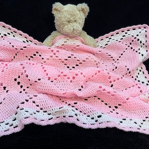 May include: A pink and white crocheted baby blanket with a teddy bear lying on top.