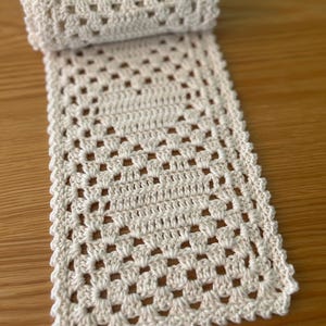 May include: A cream-colored rectangular crochet runner featuring a classic granny square stitch pattern with a decorative scalloped edge. The textile is partially rolled at one end, showcasing the intricate openwork design and consistent yarn texture.