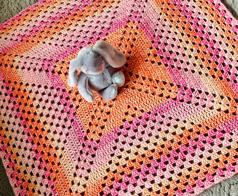 Pode incluir: Um cobertor de croch&ecirc; feito &agrave; m&atilde;o em tons de rosa e laranja, com um padr&atilde;o de granny square. Um coelho de pel&uacute;cia cinza com uma fita azul est&aacute; sobre o cobertor. O cobertor &eacute; um item macio e aconchegante.