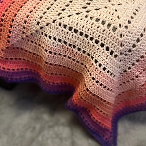 May include: A square crochet blanket featuring a gradient color pattern that transitions from a cream center to shades of pink, coral, and orange, ending in a deep purple border. The textile displays an openwork stitch design throughout.