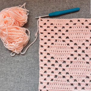 May include: A ball of light pink yarn sits next to a partially crocheted square piece of light pink fabric. A blue crochet hook is inserted into the top edge of the fabric, which features a repeating openwork pattern of stitches.