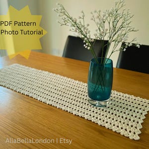 Pode incluir: Um caminho de mesa de crochê creme com um padrão geométrico está sobre uma mesa de madeira. Um vaso azul-petróleo com flores brancas está centrado no caminho de mesa. A imagem inclui o texto "PDF Pattern & Photo Tutorial".