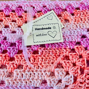 Pode incluir: Um cobertor de croch&ecirc; feito &agrave; m&atilde;o em tons de rosa e laranja, com um padr&atilde;o vazado repetido. Uma etiqueta com as palavras "Handmade with love" &eacute; presa com uma fita transparente. O cobertor &eacute; um item artesanal e aconchegante.