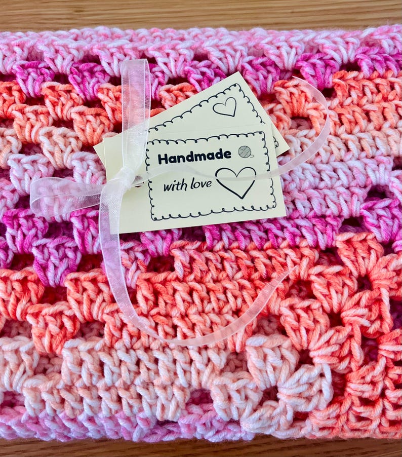 Pode incluir: Um cobertor de croch&ecirc; feito &agrave; m&atilde;o em tons de rosa e laranja, com um padr&atilde;o vazado repetido. Uma etiqueta com as palavras "Handmade with love" &eacute; presa com uma fita transparente. O cobertor &eacute; um item artesanal e aconchegante.