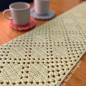 May include: A light green crocheted table runner featuring an intricate diamond pattern and openwork stitches. The text Alla Bella Designs is printed in the bottom right corner. Two ceramic mugs sit on small crocheted coasters nearby.