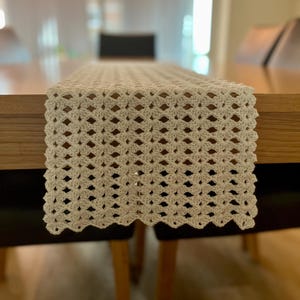 May include: A cream-coloured, crocheted table runner with a diamond pattern. The runner is displayed on a wooden table, highlighting its detailed openwork design. The edges have a scalloped finish, adding a decorative touch to the dining table.