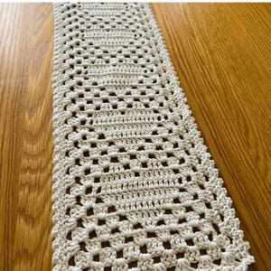 May include: Cream-colored, rectangular crochet table runner. The runner features an open, geometric pattern with a scalloped edge. The handmade textile is displayed on a wooden surface. The Etsy shop name is visible in the bottom right corner.