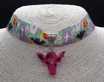 May include: A Halloween-themed choker necklace. The ribbon band features a pattern of pumpkins, bats, tombstones, and potion bottles on a teal background. A pink bat-shaped pendant with dark purple speckles hangs from the center.