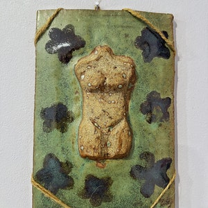 May include: A ceramic tile with a raised, textured sculpture of a torso. The tile is green with black floral designs and is tied with twine.