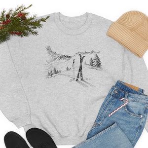 Op de afbeelding: Een grijs sweatshirt met een zwart-witte illustratie van ski's in een besneeuwd berglandschap. Het sweatshirt wordt gecombineerd met een blauwe jeans en zwarte sneakers.