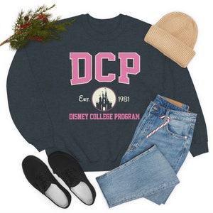 Könnte beinhalten: Ein dunkelblauer Pullover mit einem pinkfarbenen "DCP"-Logo, einem weißen Kreis mit einer Silhouette von Schloss Aschenputtel und dem Text "Est. 1981" und "DISNEY COLLEGE PROGRAM".