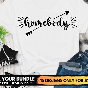 Puede incluir: Camiseta blanca con texto negro que dice "homebody" con una flecha apuntando hacia la derecha. El texto está en una fuente estilizada. La camiseta está sobre una superficie de madera con un par de zapatillas negras y unos vaqueros azules.