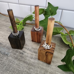 May include: Three wooden ring holders with different finishes. The holders are made of dark brown wood and have a square base. Each holder has a single wooden post that rises from the center of the base. The posts are topped with rings.