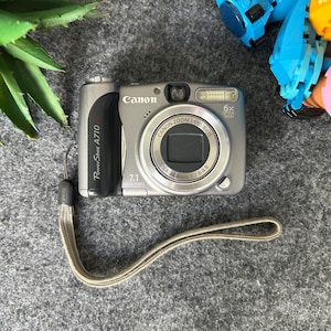 May include: A silver Canon PowerShot A710 digital camera with a 6x optical zoom lens. The camera has a 7.1-megapixel resolution and a wrist strap. The camera's lens has the following measurements: 5.8-34.8mm 1:2.8-4.8.