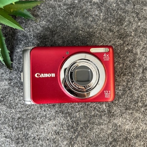 May include: A red Canon digital camera featuring a 4x optical zoom lens, 12.1 megapixels, and a silver metallic trim. The front of the camera displays the Canon logo, lens specifications of 6.2-24.8mm 1:2.7-5.6, and a built-in flash unit.