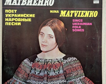 May include: Vintage vinyl record album cover featuring Nina Matviyenko. The album cover has text in both English and Ukrainian. The image shows a woman with long brown hair wearing a colorful embroidered top. The background is dark.