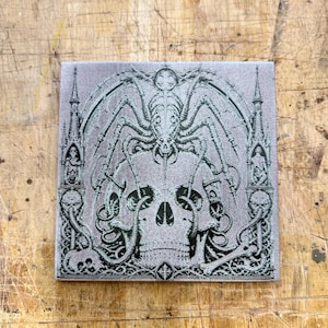 May include: Square, gray tile with a detailed black illustration of a skull, spider-like creature, and gothic architectural elements. The design is intricate and symmetrical, with a focus on dark, gothic themes.