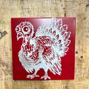 May include: A red square tile featuring a detailed white illustration of a turkey. The turkey has large eyes, a prominent wattle, and detailed feathering. The design is a stylized, artistic representation of the bird.