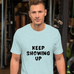 Könnte beinhalten: Ein hellblaues T-Shirt mit dem Text "KEEP SHOWING UP" in schwarzen Buchstaben. Das Shirt wird von einer Person in einem Fitnessstudio getragen.
